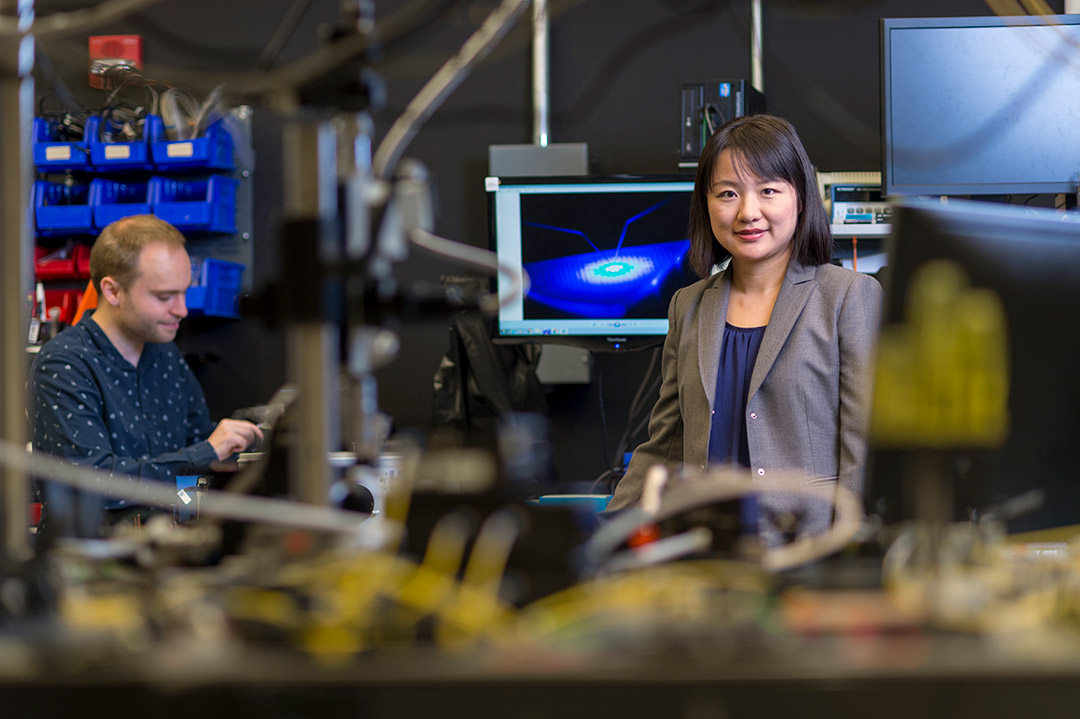 Jing Zhang is shown in the foreground, sitting in a research lab. A male colleague is shown in the background.