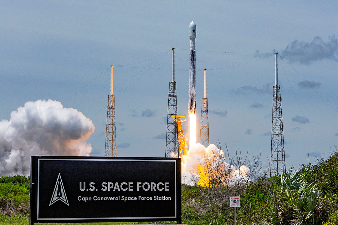 Photograph of a rocket launching from Cape Canaveral Space Force Station. The rocket is off the ground, with fire and smoke below it from launching. In the foreground, there is a black sign for the U.S. Space Force.