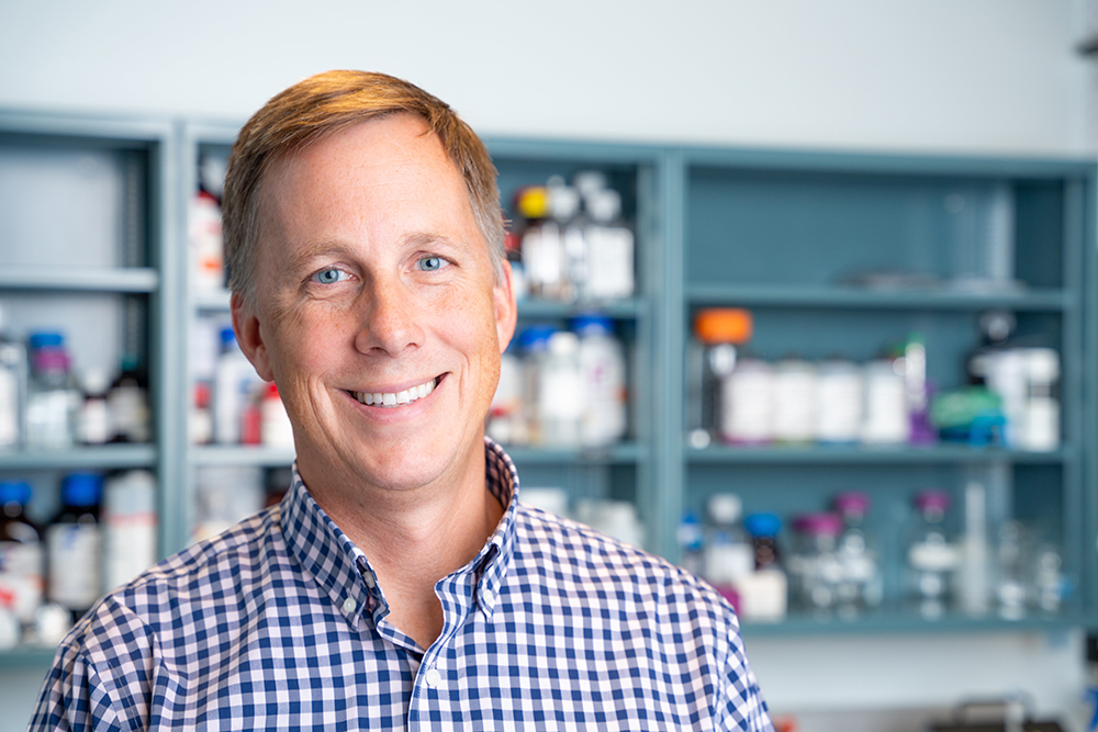 A headshot of Tom Gaborski standing in front of shelves in a lab, wearing a blue checkered button up shirt.