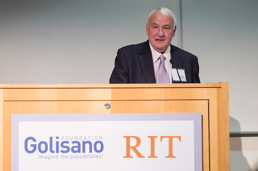 Tom Golisano stands at a podium with the RIT logo wearing a black suit and purple tie.