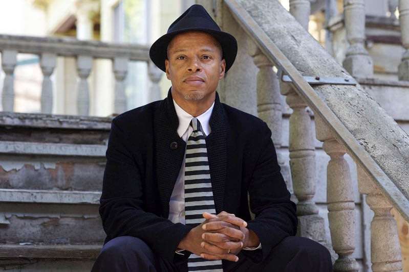 'Kerry Holiday, an middle aged african american male, sits on decorative cement steps in a black suit, black hat, and black and gray striped tie.'