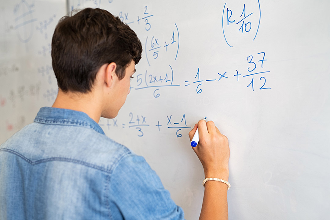 a student in a blue long sleeve shirt writes a math equation on a whiteboard.