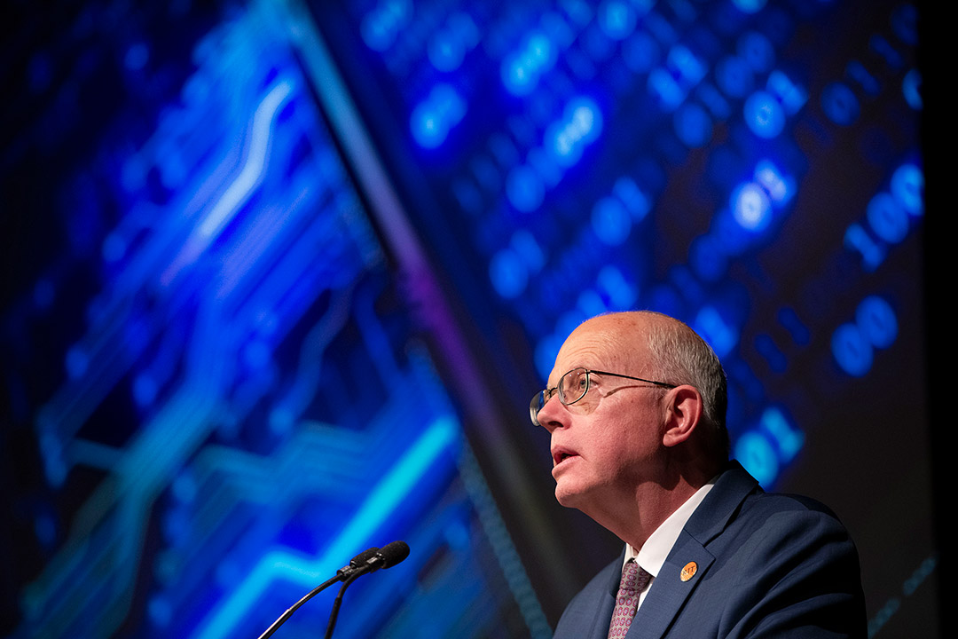 'R I T President David Munson is shown in side profile from the chest up standing at a podium. Behind him, a screen is lit up with various shares of blue resembling a computer chip.'