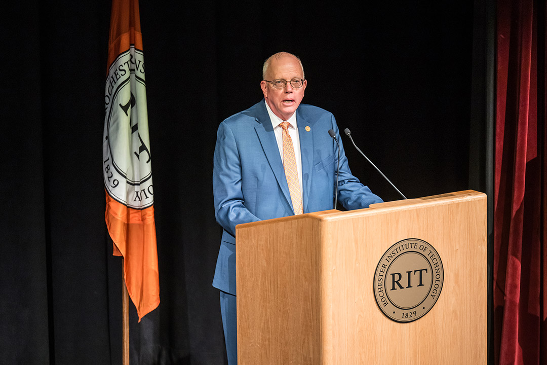 'RIT President David Munson stands in front of a podium with the RIT seal on stage.'