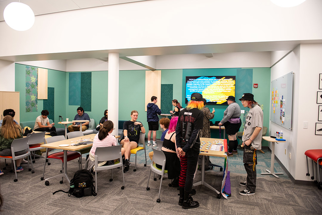 students sit at tables and stand around in a room newly renovated for meeting use.