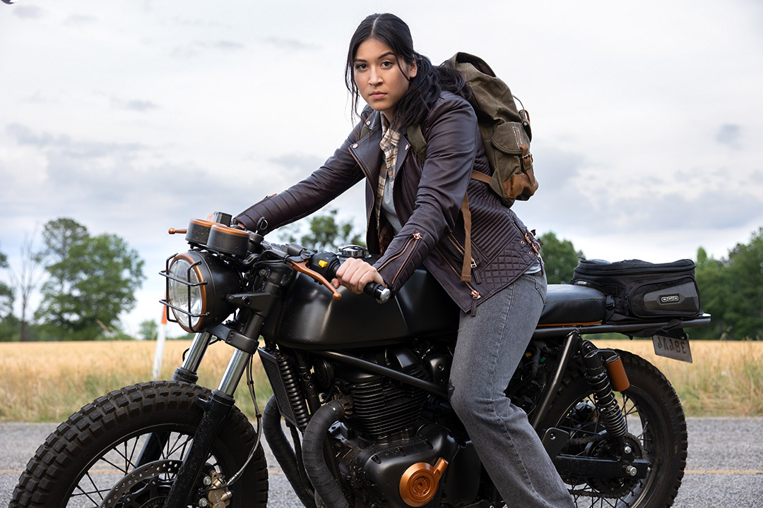 a young woman with dark hair sits on a black motorcycle wearing gray clothing and an army green backpack.