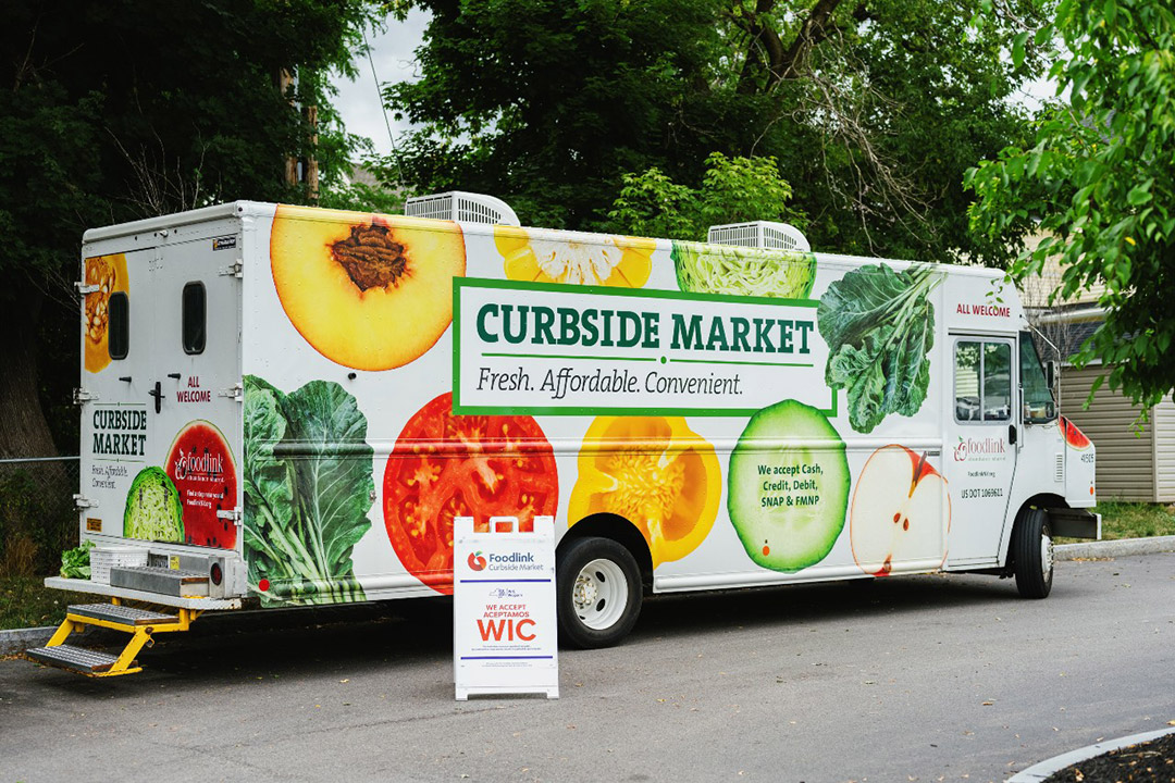 'a white truck covered in colorful pictures of fruits and veggies and bearing the logo for the Curbside Market is shown parked on a treelined street.'