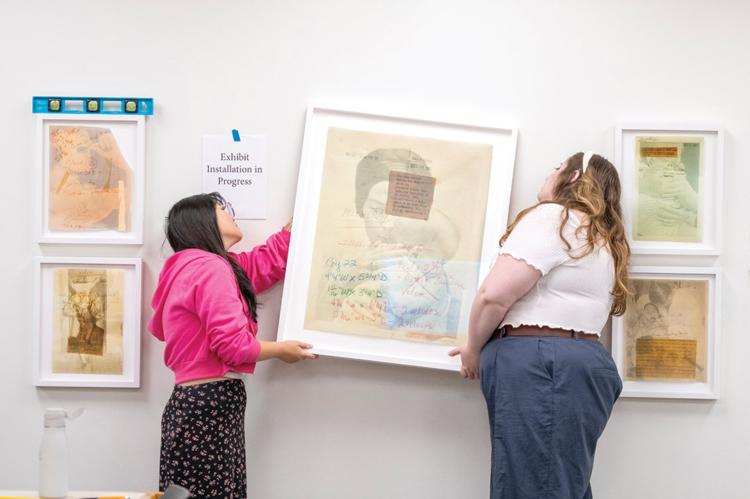 two students work together to place a large portrait on the wall of a gallery. Other smaller portraits surround them, already on the wall.