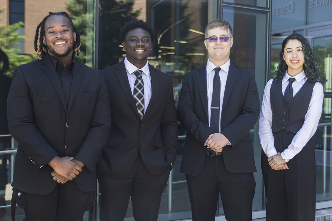 'Four students stand side by side in front of an R I T building, all wearing suits and ties.'