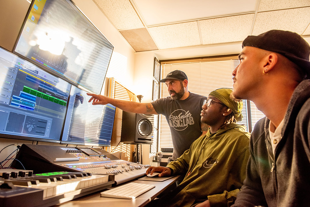 three men sit at a table with a keyboard and control panel on it. One of the men points to something on a computer screen in front of them.