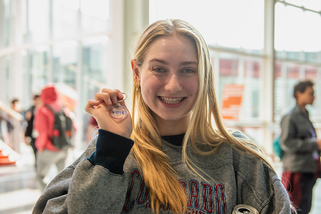 a young blonse student wearing a gray sweatshirt smiles and holds up a round clear keychain that says VOTE.