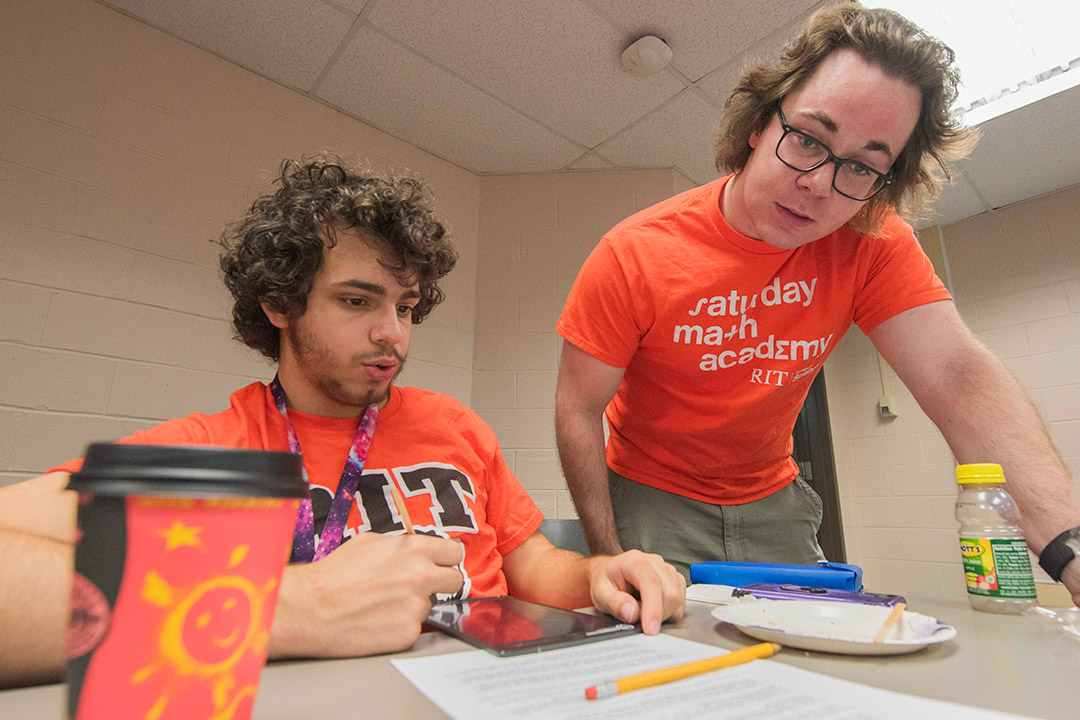 two students in orange tshirts are shown at a table with paper and a pencil in front of them. One student is actively showing the other how to do a math problem.