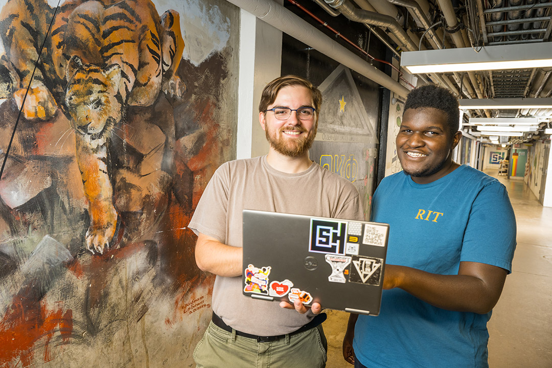 'a man in a brown tshirt and a man in a blue tshirt hold a laptop together in front of a mural of a tiger in the R I T Tunnel system.'