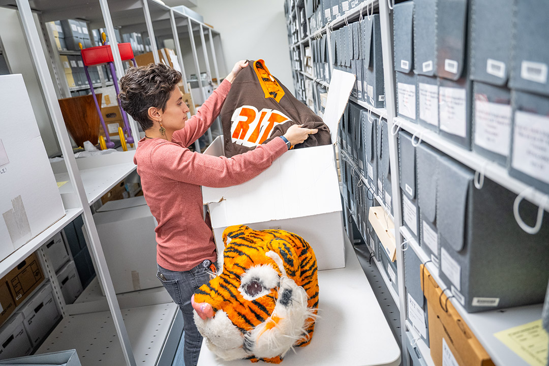 a woman with short dark hair pulls a vintage R I T jersey and mascot head out of a white box in an archive repository setting.