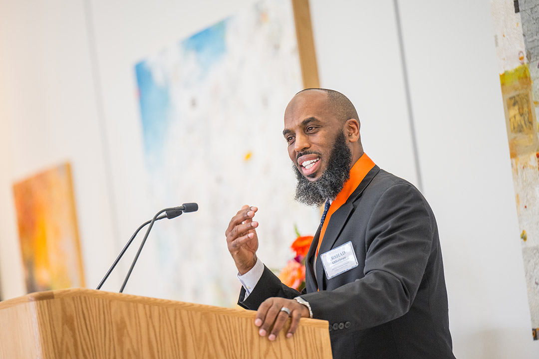 a black man with a beard stands at a podium wearing a black suit jacket with an orange dress shirt, addressing an audience.