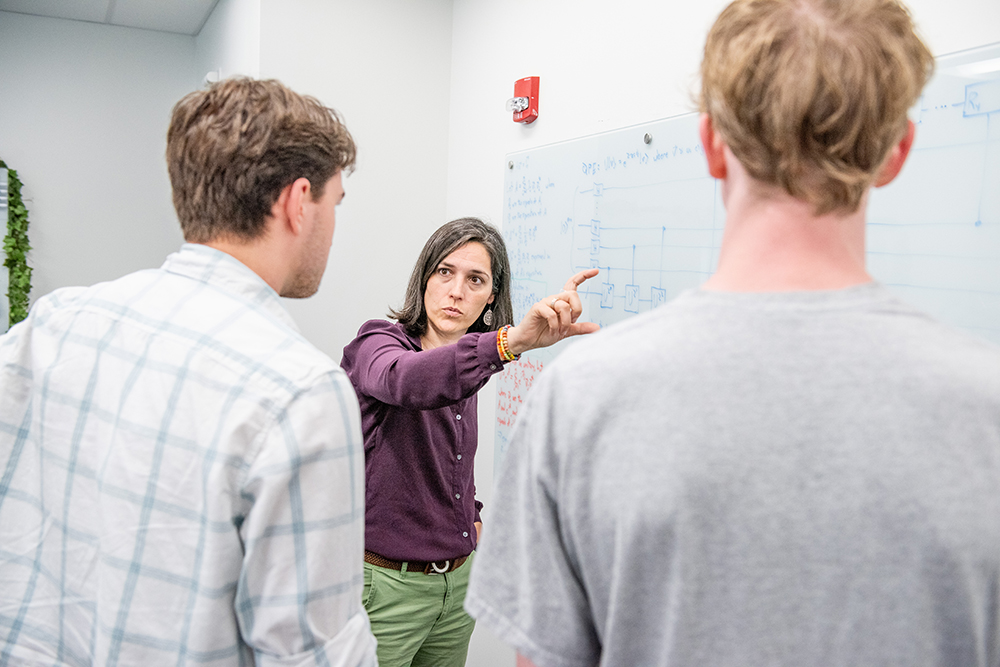 A professor standing between two students and in front of a white board, explaining something to the students.