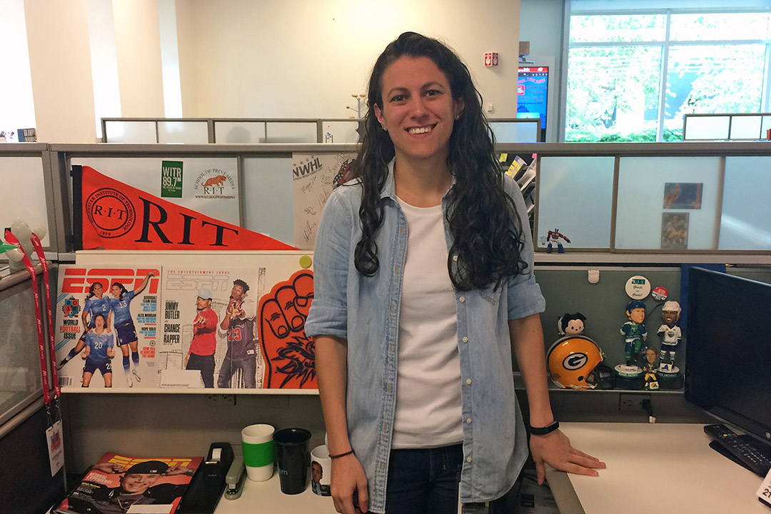 a woman standing at her desk, decorated with E S P N magazines and R I T gear.