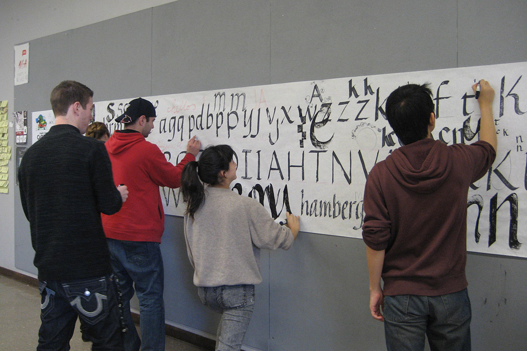 students draw large calligraphy letters on a poster on a wall.