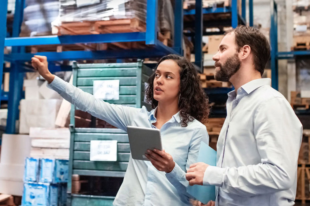 Two people stand in a warehouse looking at stocked shelves and a tablet computer.