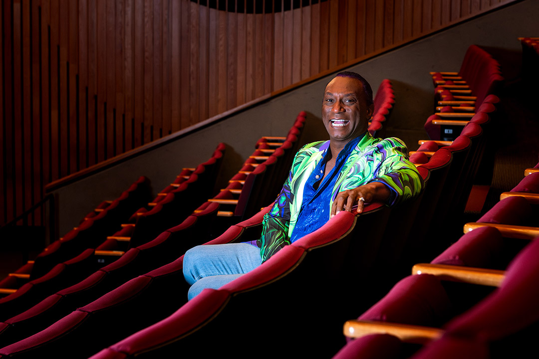 'Thomas Warfield sitting in a theater seat with rows of seats all around. Thomas is wearing a colorful jacket, bright blue shirt, light blue pants. '