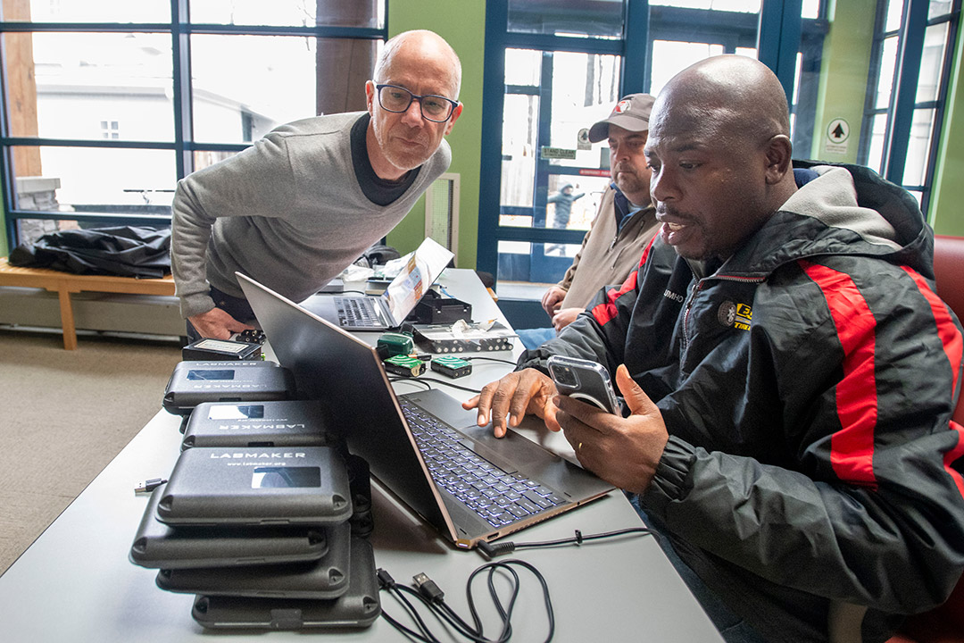 two men sit at a table looking at a laptop. A third man leans over the table to look at the laptops screen.