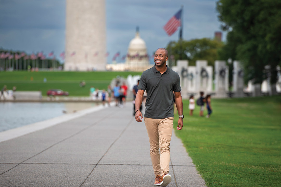 Arthur Deane walks through a park like setting in front of the capitol building wearing a dark polo shirt and khaki pants.