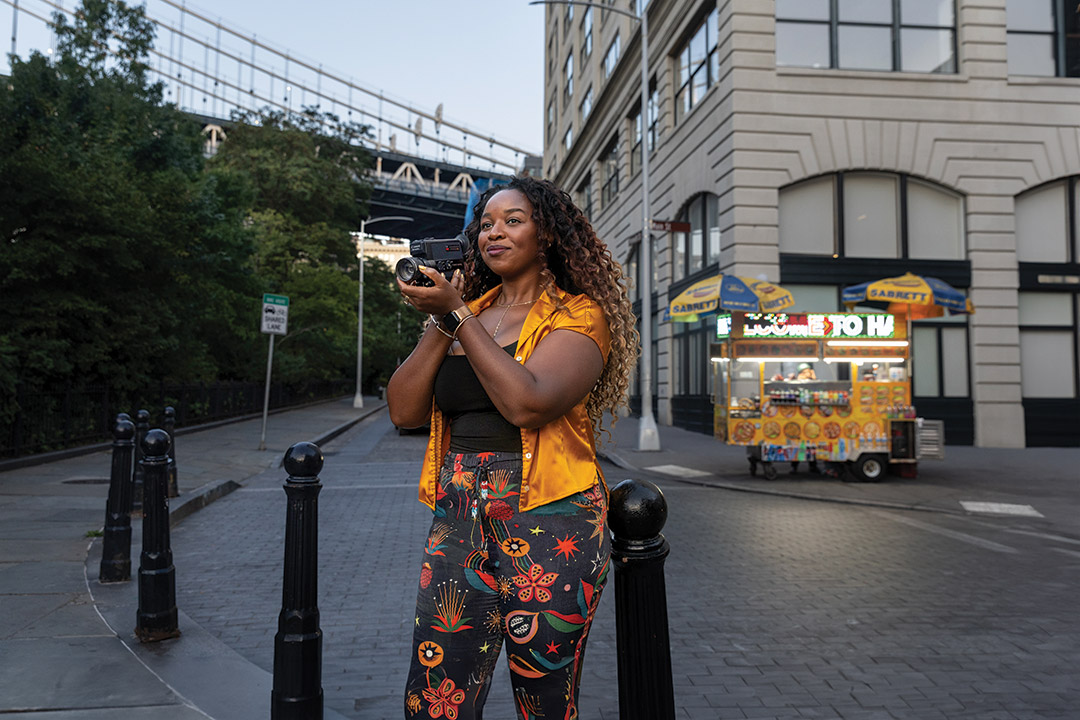 Chris Wairegi stands on a street corner in New York City with a smile on her face and hands clasped near her face.