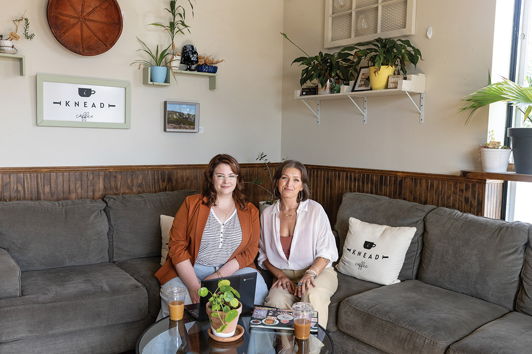 two women sit on a gray couch in a white livingroom with art on the walls.