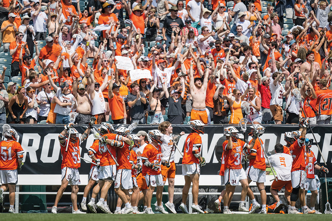 The R I T lacrosse team celebrates on a turf field in front of a crowd of people.