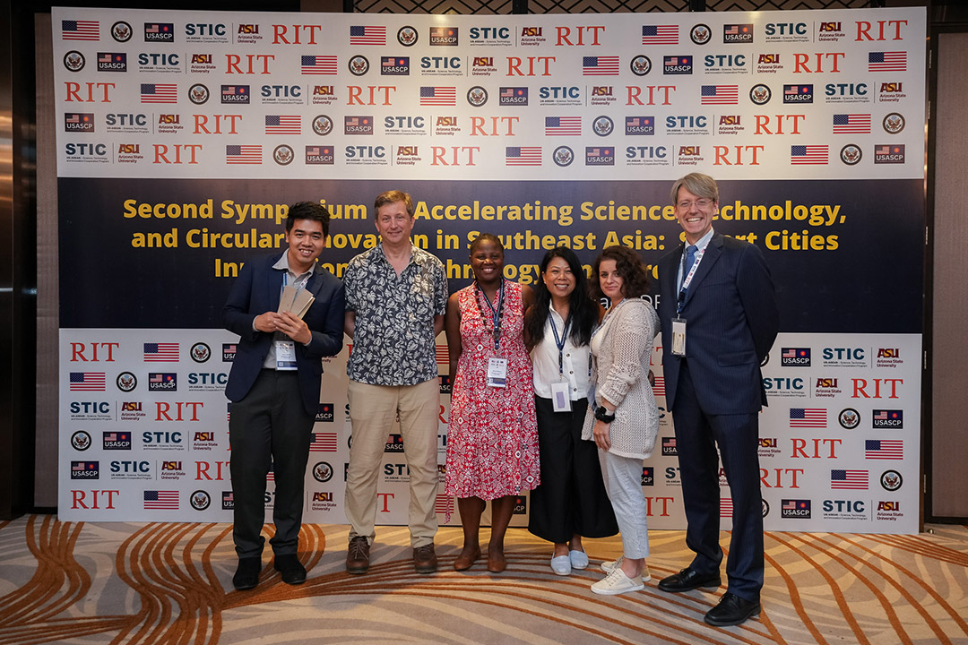 'a group of six students and adults stand in front of a sign bearing logos from R I T and other companies.'