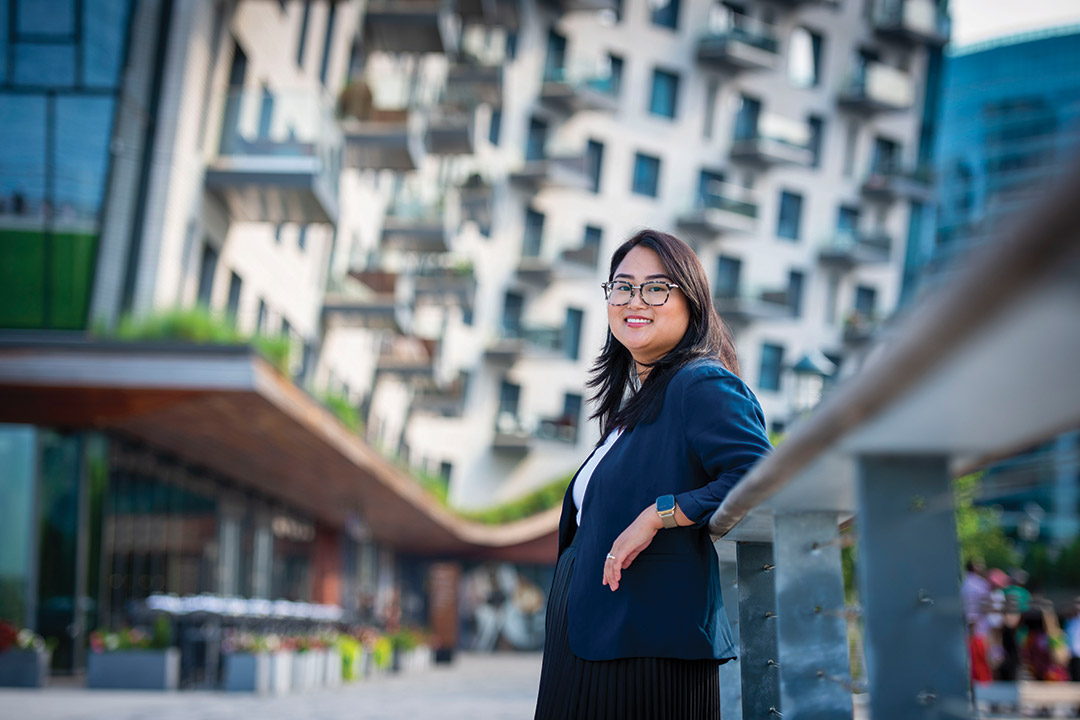 Valerie Horn wears a blue suit jacket and leans against a railing in an outdoor area surrounded by tall buildings.