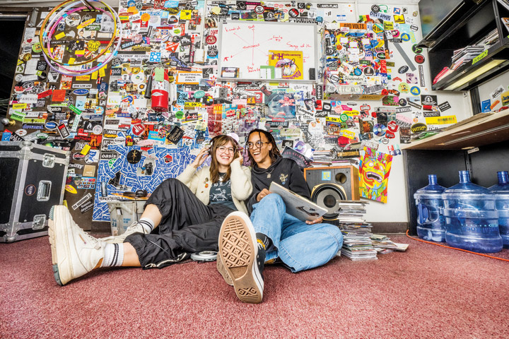 Two students sit on the floor of a radio booth wearing headsets.