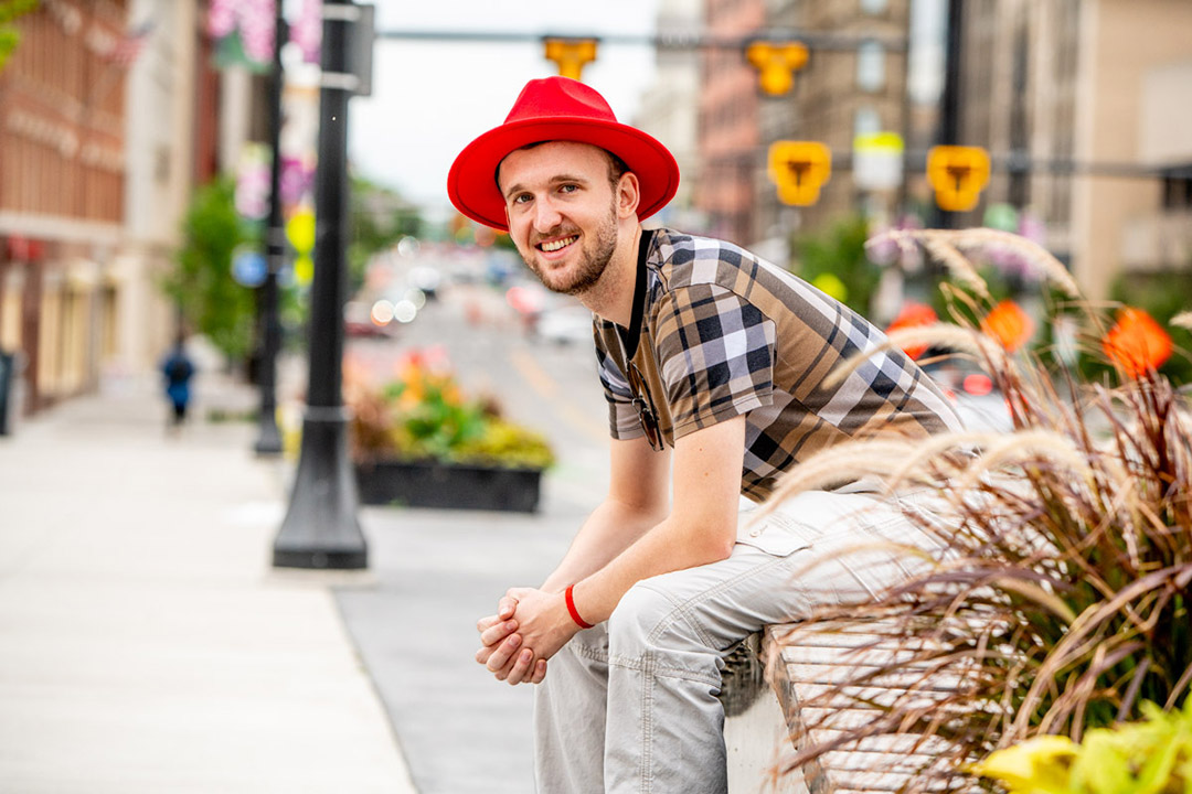 a man in a red fedora and plaid shirt sits on a curb in a city setting.