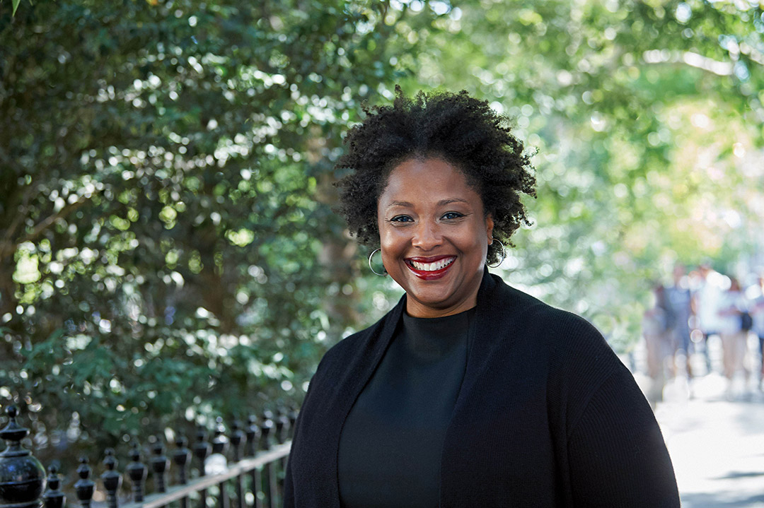 'an older black woman stands in a park like setting with black clothing and a big smile.'