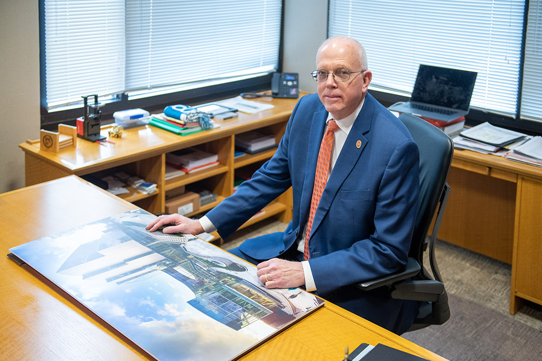 David Munson wears a blue suit and sits at a desk in his office with a large image of R I T's shed building in front of him.