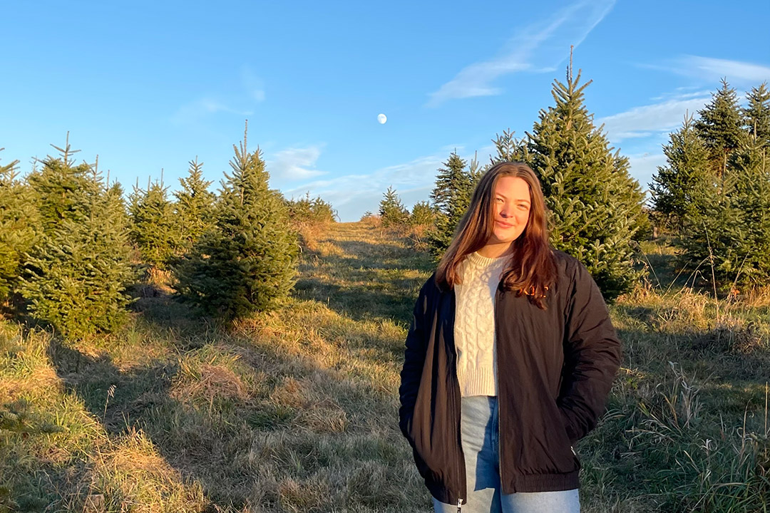 'a college age girl standsin a black jacket and cream sweater in a forest area.'