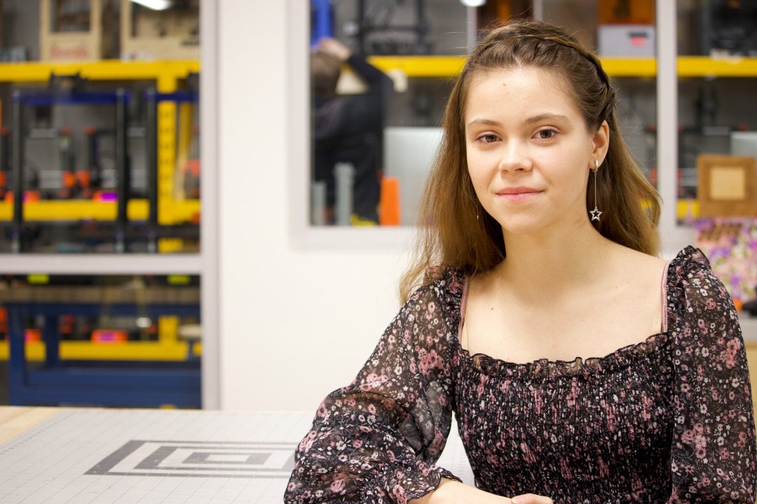 a college age girl in a black floral dress sits at a table in a lab room towards the right side of the image