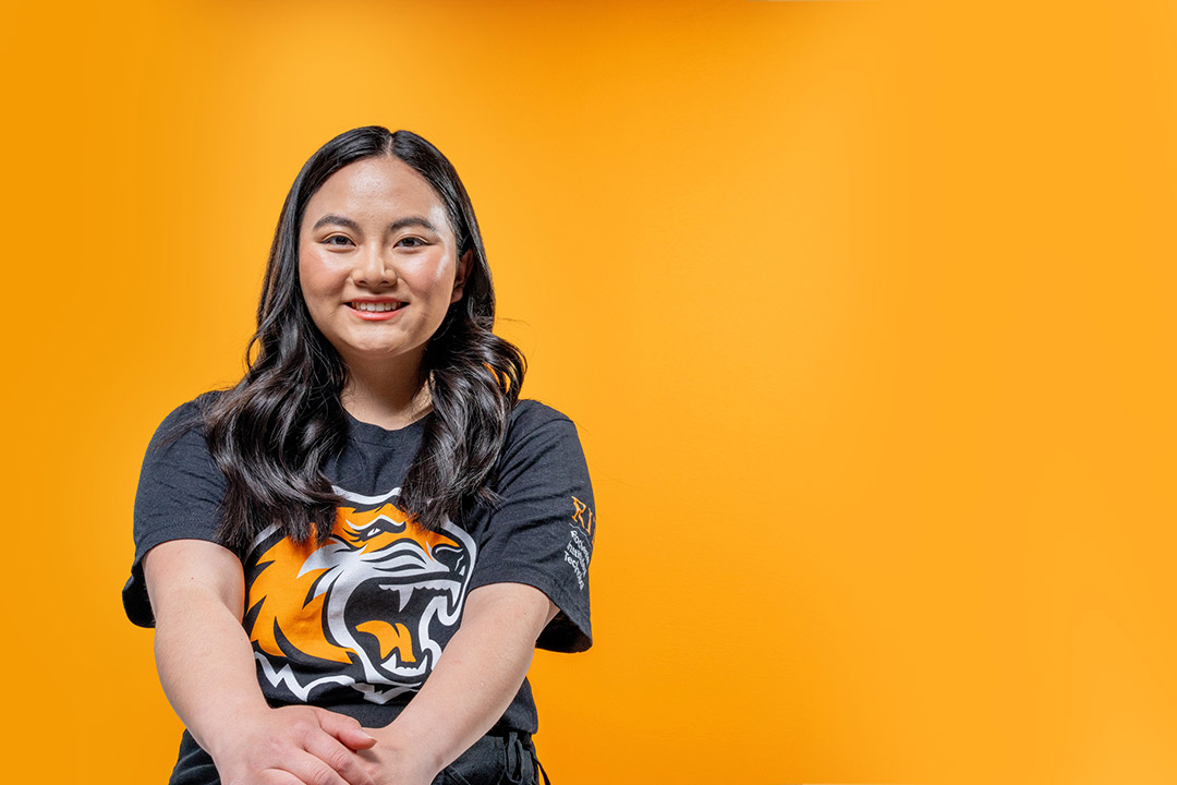 Emma Kane, a young college student, sits in a black shirt with a tiger on it in front of an orange background.