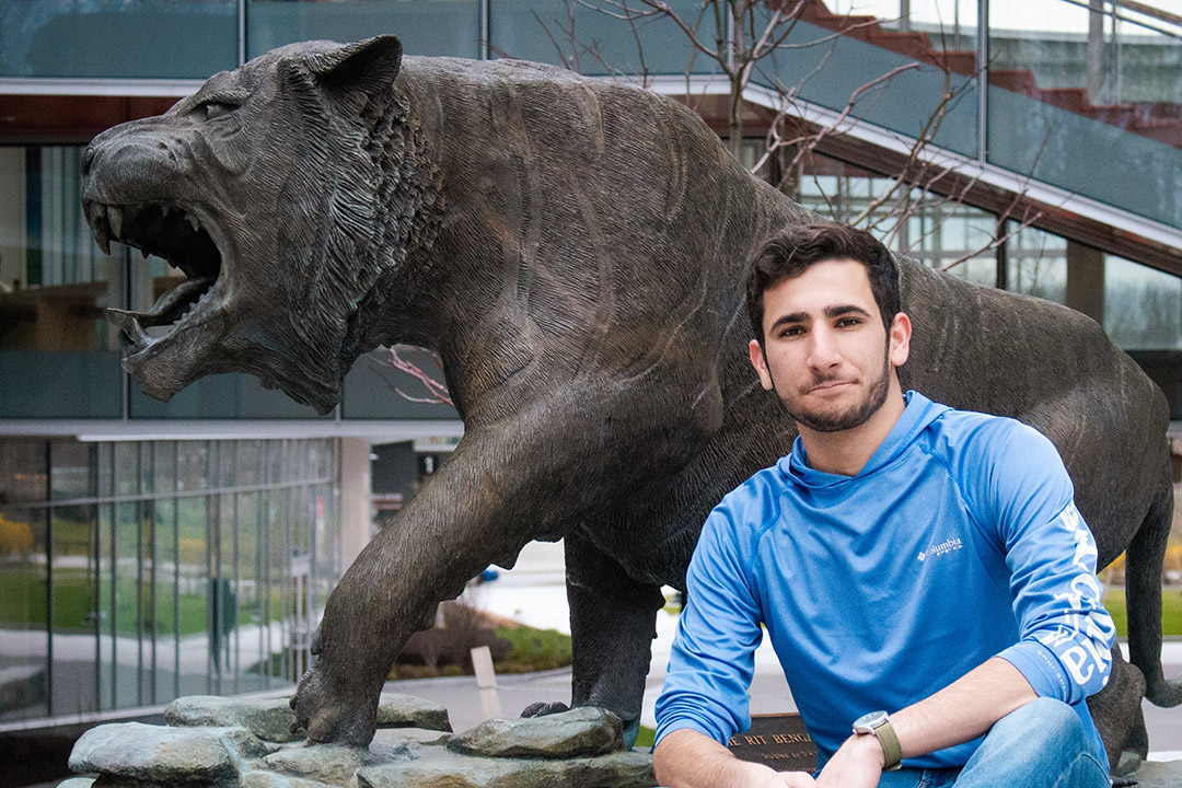 'Kyle Scher kneels in front of a tiger statue on campus wearing a long sleeve blue shirt.'