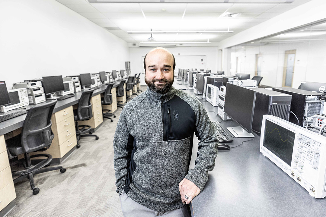 a man in a gray long sleeve shirt stands with his arm resting on a countertop in a computer lab.