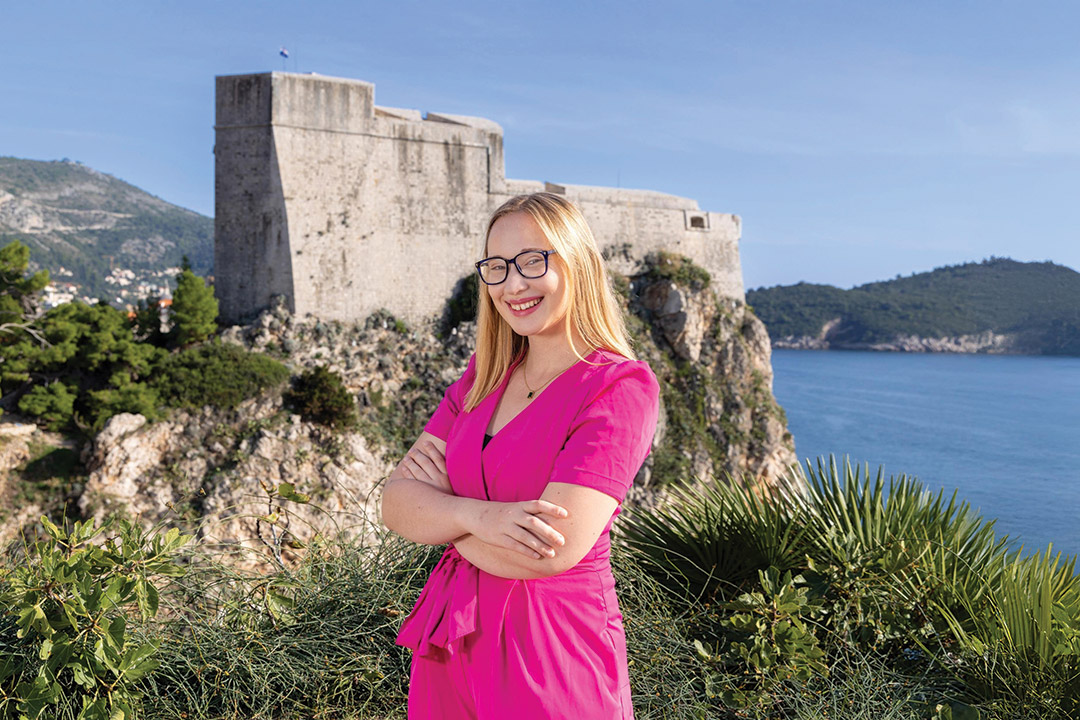 a college age girl in a pink dress stands on a Croatian coastline.