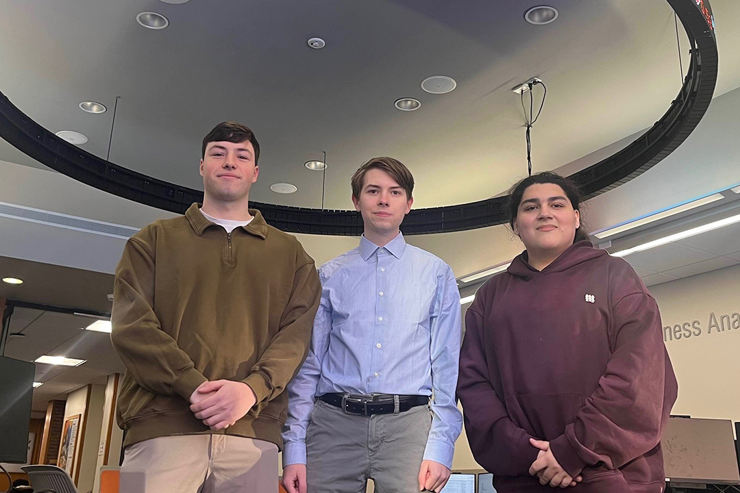 Three students stand under a bloomberg trading terminal hanging from the ceiling.