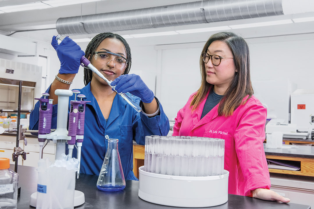 a college age girl holds a pipette in a lab while a faculty member looks on.