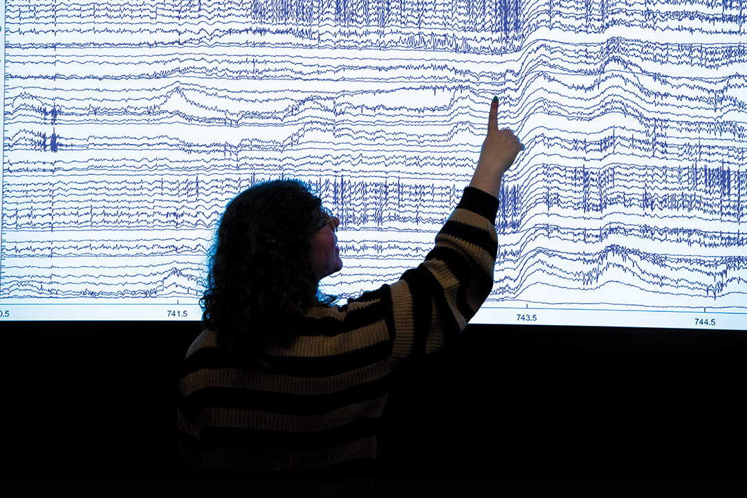 The silouhette of a college age girl is shown pointing at a large screen with brain waves showing.