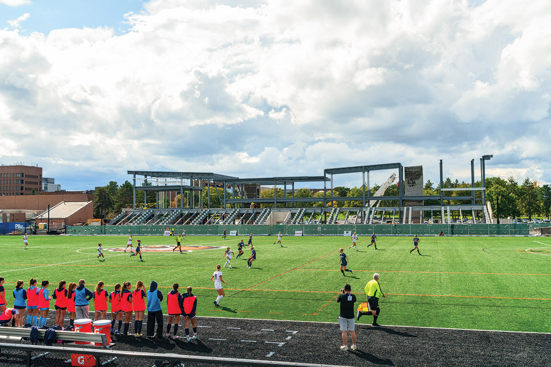 students are shown playing on the turf of a field with a half built stadium in the background.