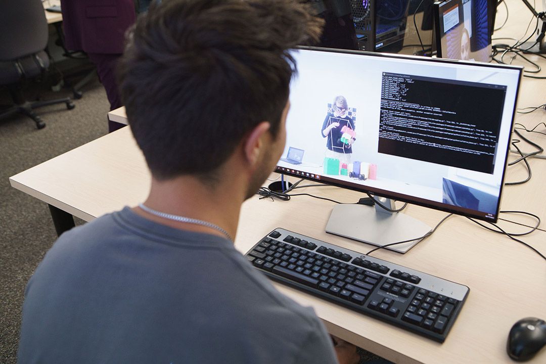 A person with short dark hair sits at a desk, facing a computer monitor displaying a coding interface and a video feed of someone handling colored blocks. A keyboard and mouse are on the desk, and additional monitors and office furniture are visible in the background.