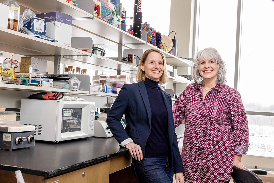 Two smiling women stand in a well-equipped laboratory, surrounded by scientific equipment, shelves of supplies, and colorful molecular models.