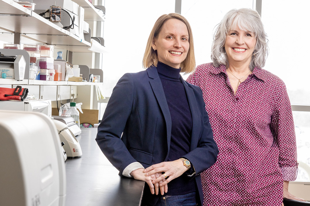 Two smiling women stand in a well-equipped laboratory, surrounded by scientific equipment, shelves of supplies, and colorful molecular models.