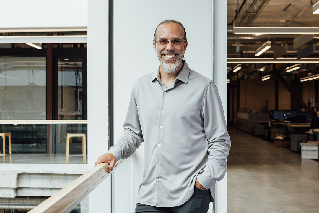 Smiling man with glasses and a beard, wearing a light gray button-up shirt, leans on a railing in a modern office space.