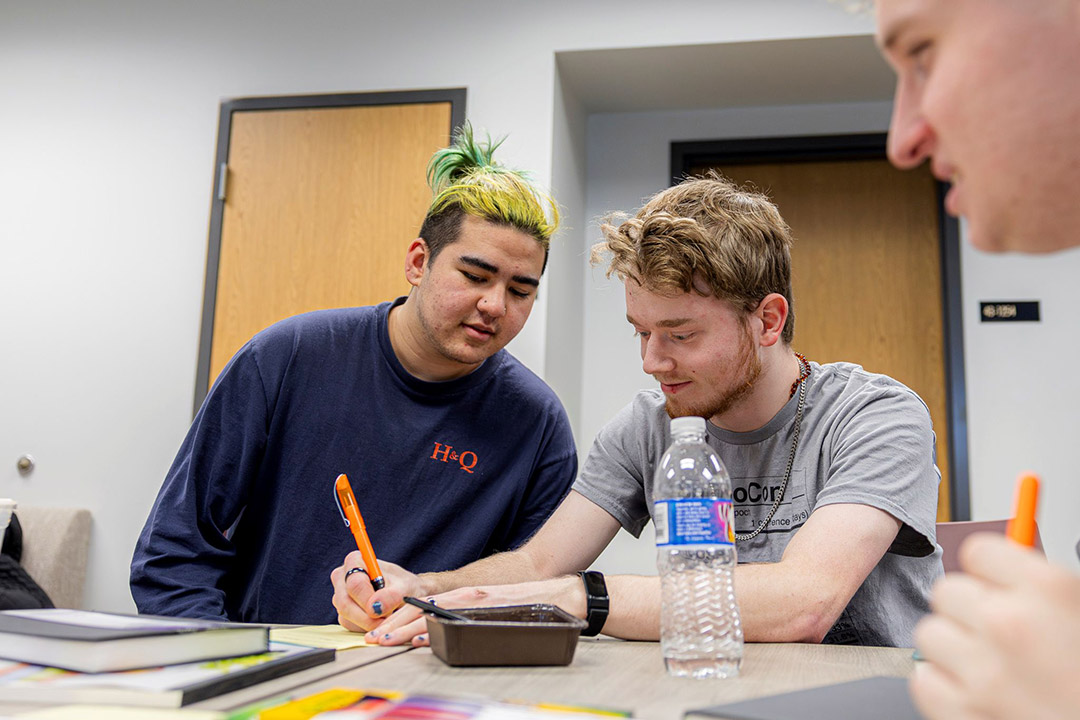 Two students collaborate at a table in a classroom, one holding a pen and writing while the other leans in to assist. Books, papers, and a water bottle are on the table, with other students in the background.